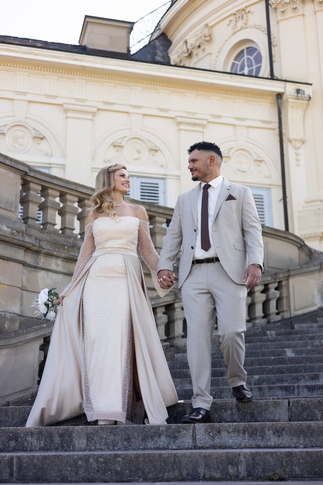 Paar geht Hand in Hand eine Schlosstreppe hinunter, sie im Perlenkleid, er im hellen Anzug.