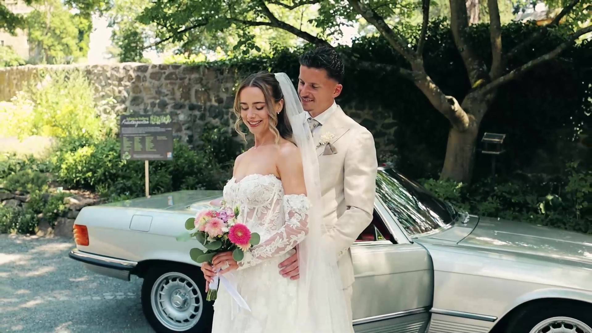 Standbild aus einem Hochzeitstrailer: Brautpaar vor einem silbernen Oldtimer.